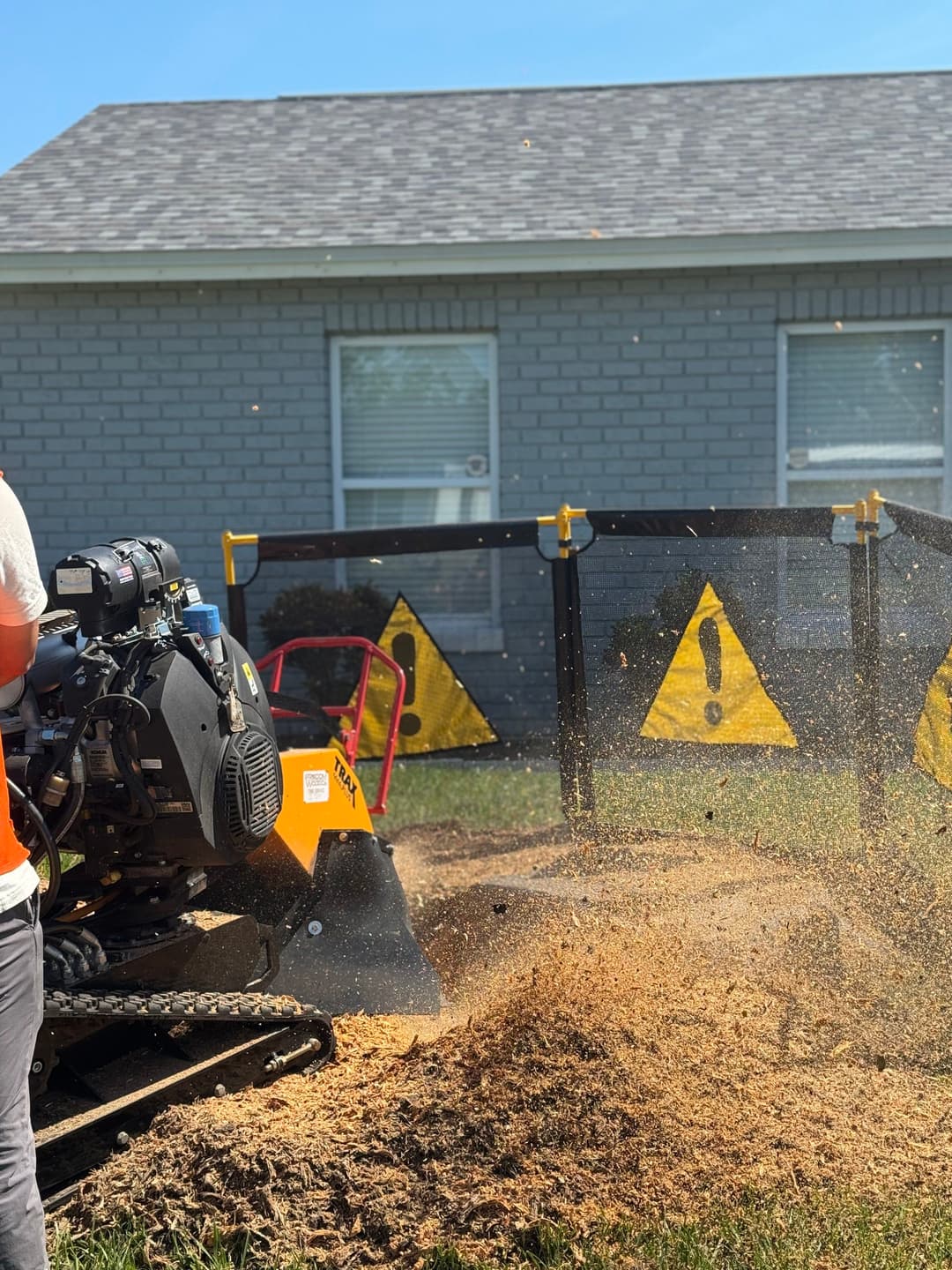 Tree stump removal with a stump grinder near a house, surrounded by safety caution signs.