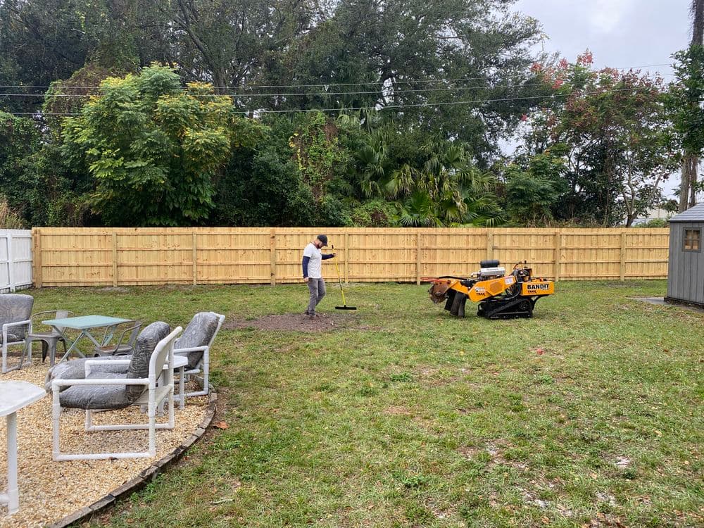 Person using a mini excavator in a backyard with a wooden fence and outdoor furniture.